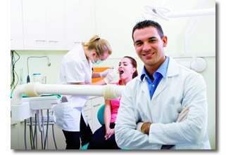 Dentist smiling with patient in the background getting their teeth checked out.